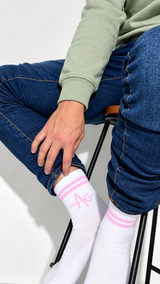 White crew socks with pink stripes and a pink monogram logo, worn by a model sitting on a stool in blue jeans and a sage green sweatshirt.