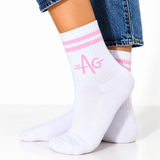 White crew socks with pink stripes and a pink 'THE AG' logo, worn on a person's feet with blue jeans visible at the top.