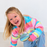 A young blonde girl smiling and laughing while kneeling on a light floor, wearing a pastel rainbow striped long-sleeve crewneck sweatshirt with The AG logo and blue jeans, holding a small round magnifying glass.