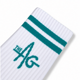 A close-up of a white ribbed sock with two teal stripes near the cuff and a large teal 'THE AG' logo knitted into the side, shown on a clean white background.