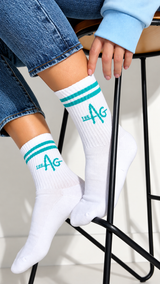 White crew socks with teal stripes and a teal 'THE AG' logo, worn by a model sitting on a black metal stool.