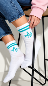 White crew socks with teal stripes and 'THE AG' logo, worn by a model sitting on a black stool.