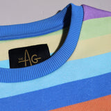 Close-up of a rainbow-striped crewneck sweatshirt by The AG, showing blue ribbed collar, black brand label with gold lettering, and pastel stripes of yellow, green, purple, blue, and orange.