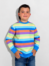 A young boy wearing a long-sleeve crewneck sweatshirt with horizontal stripes in blue, purple, orange, yellow, and green, paired with blue jeans, standing against a light gray wall.