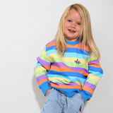 A young girl with blonde hair smiling, wearing a colorful horizontally striped sweatshirt with blue ribbed cuffs and hem, paired with light blue jeans, standing against a plain white wall.