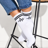 White crew socks with black stripes and 'THE AG' printed on the cuff, worn on a person's feet while sitting on a black metal stool.