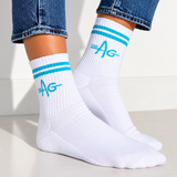 White ankle socks with two turquoise stripes and a turquoise 'THE AG' logo, worn with cuffed blue jeans on a light neutral floor.