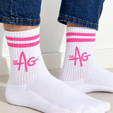 White crew socks with pink stripes and a pink 'THE AG' logo, worn with blue jeans. The socks are shown on a person's feet against a clean, light-colored background.