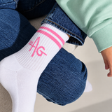A close-up of a woman's leg wearing a white crew sock with pink stripes and a pink 'AG' logo, paired with blue jeans and a mint green sweatshirt.