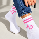 White crew socks with pink stripes and 'the AG' logo, worn with blue jeans. The brand 'the AG' is clearly visible on the side of the socks.